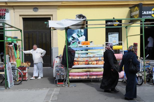 Ein Marktstand mit Stoffen, an dem zwei Frauen vorbeigehen; ein Mann steht daneben.