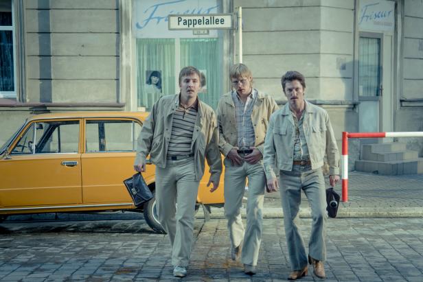 Drei Männer im Stil der 1970er Jahre gehen auf einer Straße entlang.