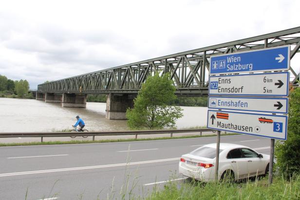 Ein Radfahrer fährt an der Enns entlang, im Hintergrund eine Eisenbahnbrücke und Wegweiser nach Wien, Enns und Mauthausen.