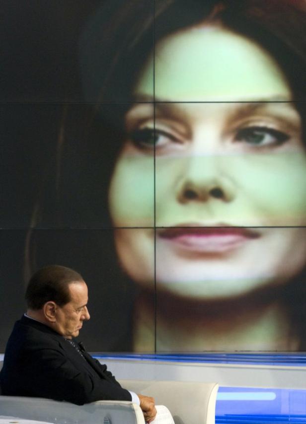 Silvio Berlusconi sitzt vor einem Bildschirm, der das Porträt einer Frau zeigt.