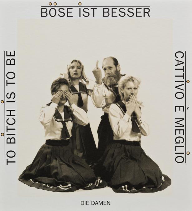 Die Gruppe „Die Damen“ in Matrosenuniformen, posiert mit den Händen vor Augen, Ohren und Mund, während ein Mann den Mittelfinger zeigt.