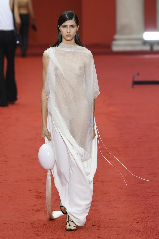 Eine Frau in einem transparenten Oberteil und einem langen weißen Rock auf einem roten Laufsteg.