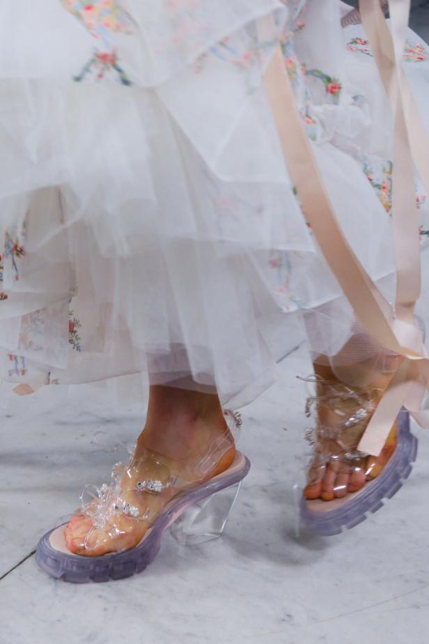 Eine Person trägt transparente Sandalen mit Absatz und ein weißes Kleid mit Blumenmuster.
