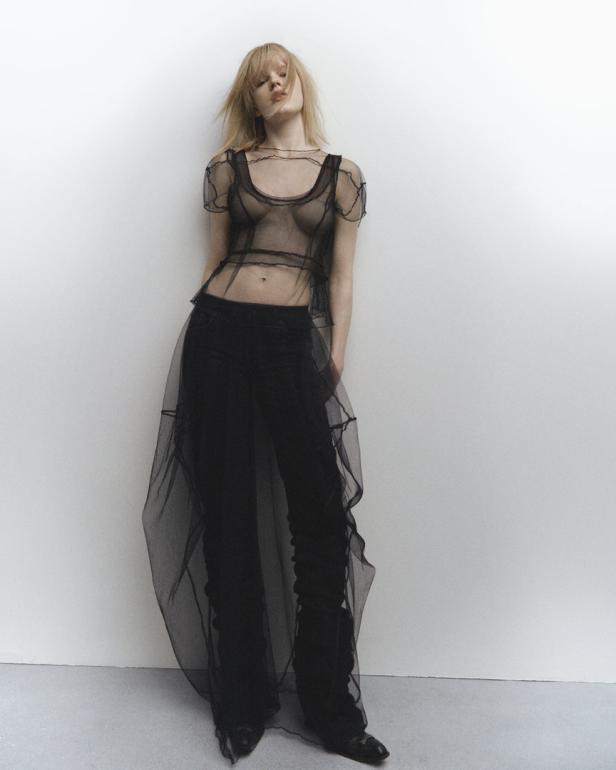 Eine Frau mit blonden Haaren posiert in einem transparenten schwarzen Oberteil und einer Hose vor einer weißen Wand.