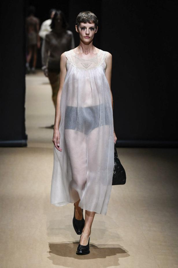 Ein Model mit dunklem Make-up trägt ein transparentes, weißes Kleid und schwarze Schuhe auf dem Laufsteg.
