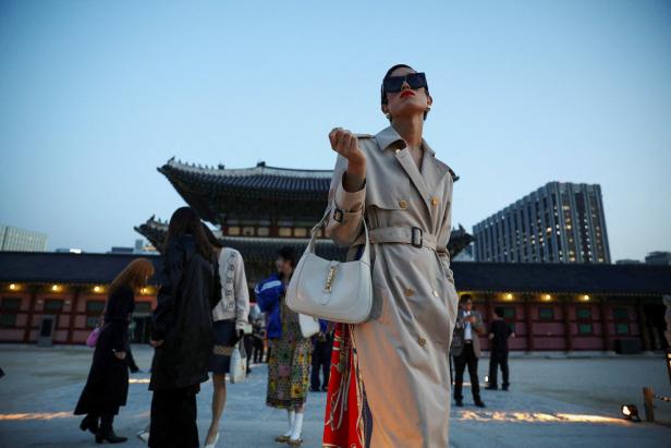 Eine Frau mit Trenchcoat und Handtasche posiert vor einem traditionellen koreanischen Gebäude.