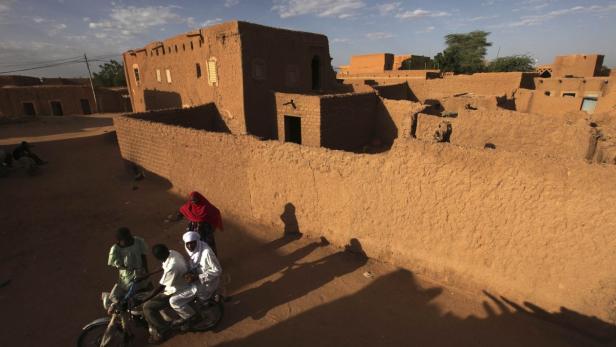Eine Gruppe von Menschen fährt mit einem Motorrad durch ein Dorf mit Lehmbauten.