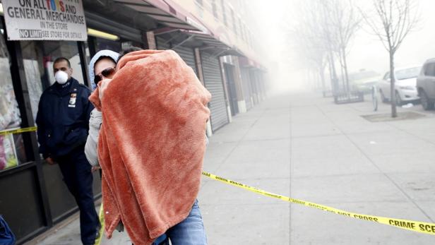 Eine Person gehüllt in eine Decke steht vor einem Laden, während ein Polizist mit Maske im Hintergrund steht.