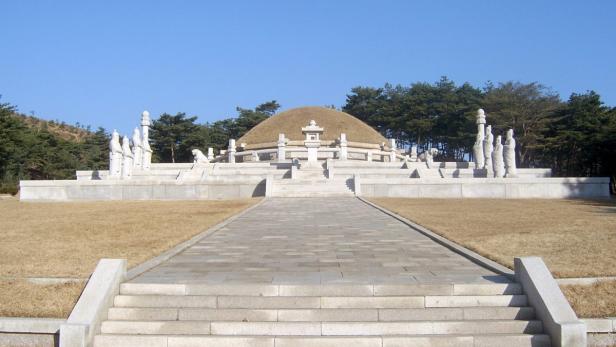 Ein Hügelgrabmal mit Statuen und einer gepflasterten Zufahrt in einer parkähnlichen Umgebung.