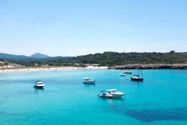Mehrere Boote ankern in einer türkisfarbenen Bucht mit einem Sandstrand im Hintergrund.