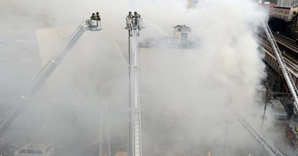 Feuerwehrleute bekämpfen einen Brand von Drehleitern aus in einer von Rauch verhüllten städtischen Umgebung.