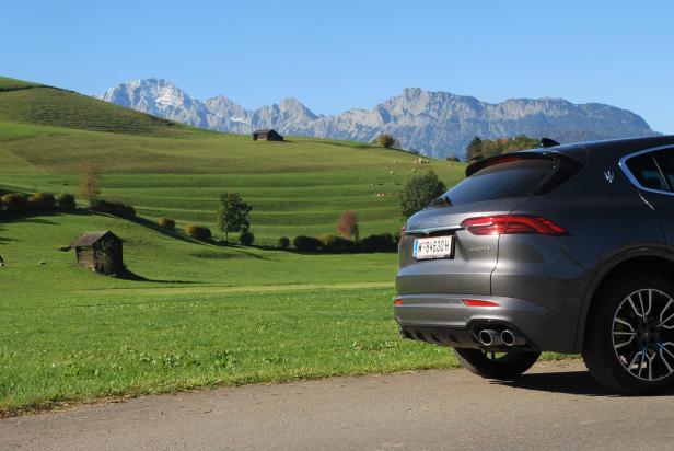 Ein grauer Maserati Levante vor einer grünen Hügellandschaft mit schneebedeckten Bergen.