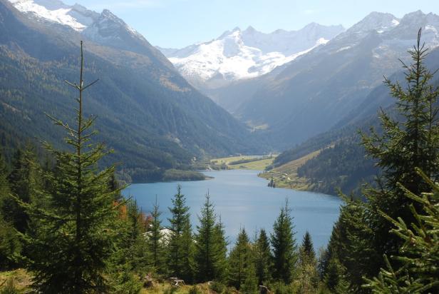 Ein Bergsee liegt eingebettet zwischen bewaldeten Hängen und schneebedeckten Gipfeln.