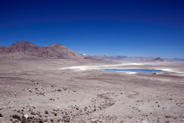 Weite, trockene Landschaft mit Bergen und einem See unter blauem Himmel.