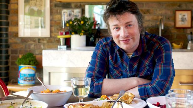 Jamie Oliver sitzt an einem Tisch mit Speisen und Getränken.
