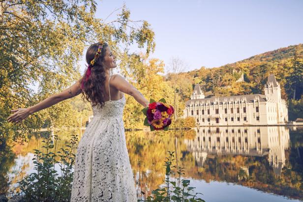 Eine Frau im Brautkleid steht mit einem Blumenstrauß vor dem Schloss am See.