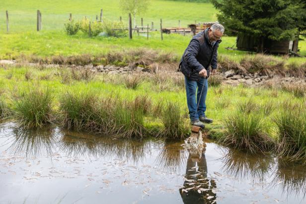 Ein Mann steht am Ufer eines kleinen Sees und stochert mit einem Stock im Wasser.