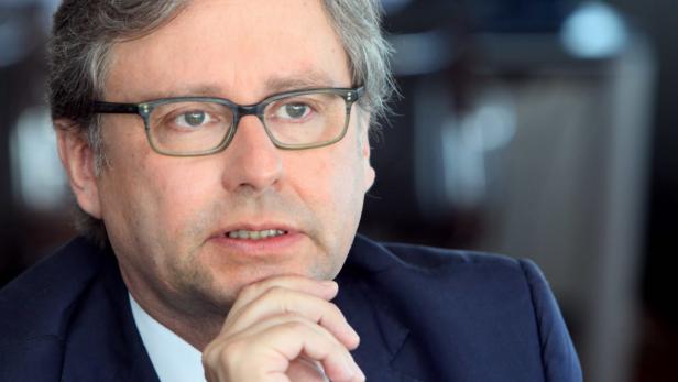 Ein Mann mit Brille und dunklem Anzug hält seine Hand ans Kinn.
