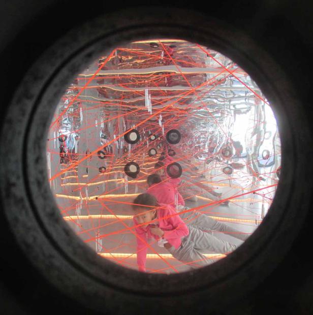 Ein Mädchen klettert durch ein Spinnennetz aus roten Laserstrahlen in einem Spiegelkabinett.