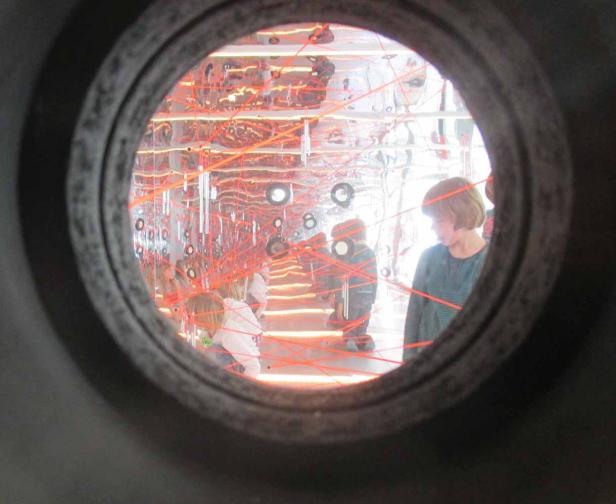 Kinder spielen in einem Raum mit verspiegelten Wänden und roten Laserstrahlen, gesehen durch ein Bullauge.