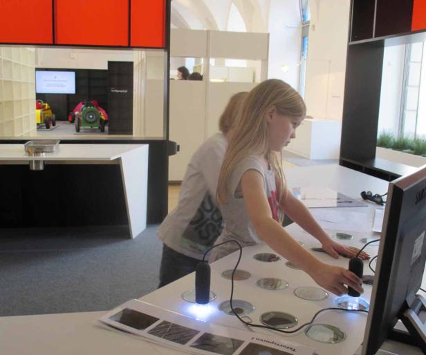 Ein Mädchen untersucht mit einem Mikroskop Proben an einem Tisch mit einem Monitor.