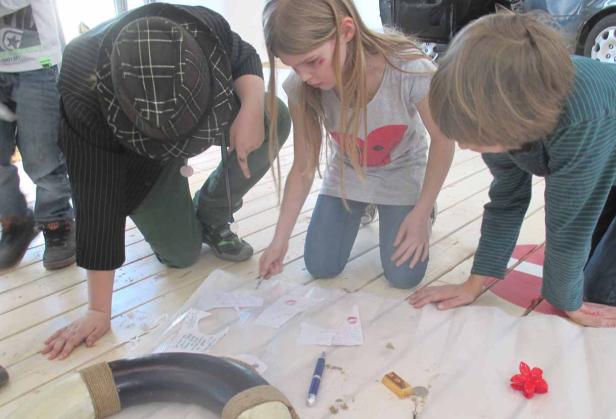 Drei Kinder knien auf dem Boden und betrachten Zettel mit Abdrücken und Zeichnungen.