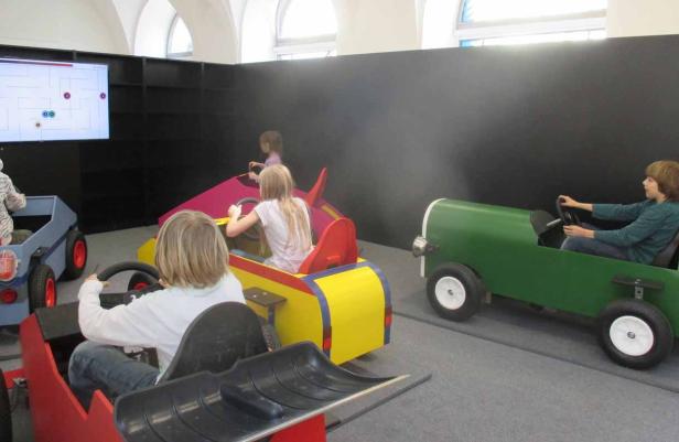 Kinder fahren in bunten Spielzeugautos vor einem Bildschirm mit einem Labyrinth.