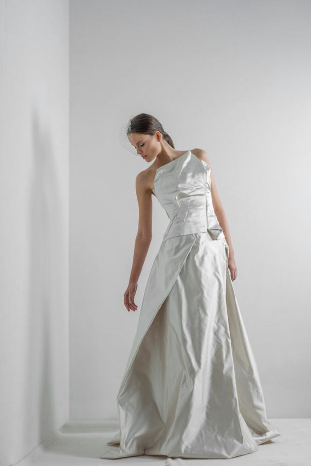 Eine Frau in einem asymmetrischen, weißen Brautkleid mit Schleier posiert vor einer weißen Wand.