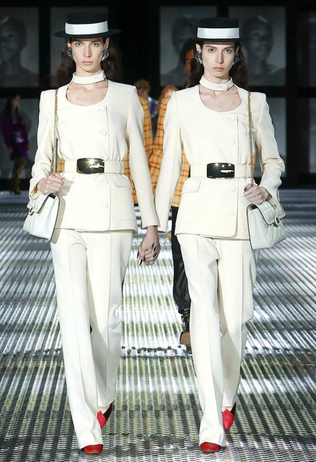Zwei Models in weißen Anzügen und Hüten laufen Händchen haltend über einen Laufsteg.