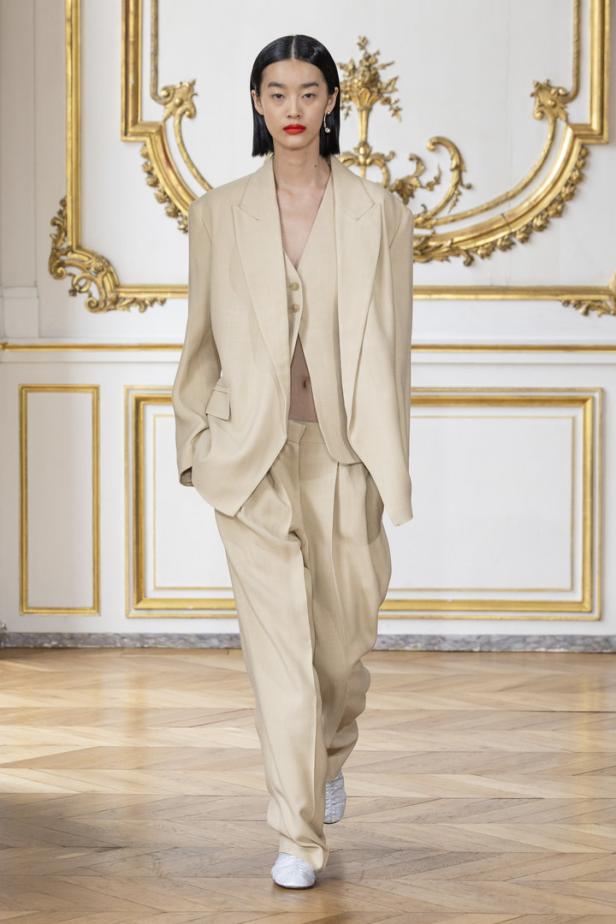 Eine Frau präsentiert ein beigefarbenes Hosenanzug mit übergroßen Blazer auf einem Laufsteg.