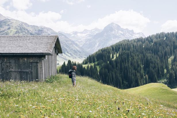 Ein Wanderer steht auf einer Blumenwiese mit einer Hütte und Bergen im Hintergrund.