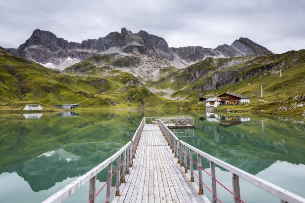 Ein Steg führt über einen Bergsee mit Blick auf grüne Hänge und schneebedeckte Gipfel.