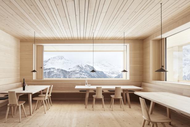 Ein holzvertäfelter Raum mit Tischen und Stühlen vor einer Fensterfront mit Blick auf schneebedeckte Berge.
