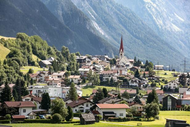 Ein Dorf mit einer Kirche mit rotem Turm liegt vor einer bewaldeten Bergkulisse.