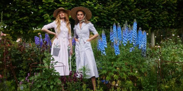 Zwei Frauen mit Strohhüten und Sommerkleidern stehen in einem blühenden Garten.