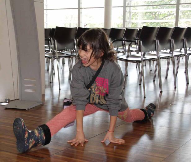 Ein Mädchen macht einen Spagat auf einem Holzboden vor einer Reihe von Stühlen.