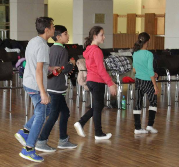 Vier Kinder gehen in einem Raum mit Stühlen an den Seiten entlang.