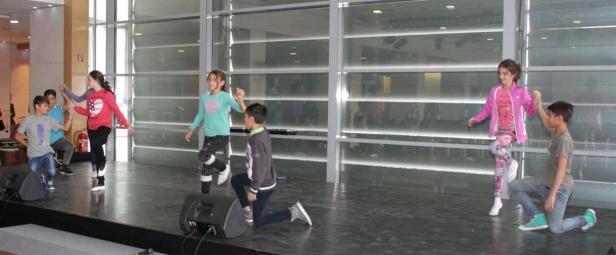 Eine Gruppe Kinder tanzt auf einer Bühne vor einem großen Fenster.