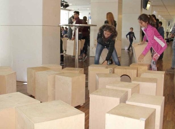 Zwei Mädchen bewegen Holzkisten in einem Raum, während andere Personen im Hintergrund stehen.
