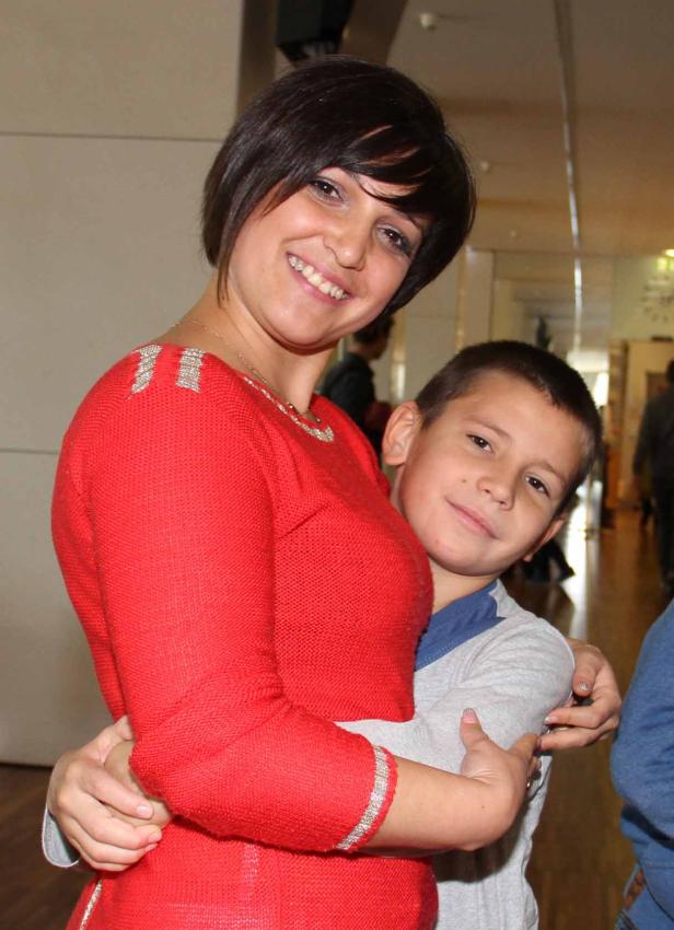 Eine Frau umarmt einen Jungen und lächelt in die Kamera.