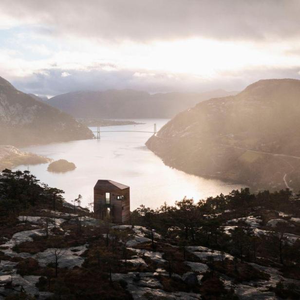 Eine kleine Holzhütte auf einem Hügel mit Blick auf einen Fjord und eine Brücke.