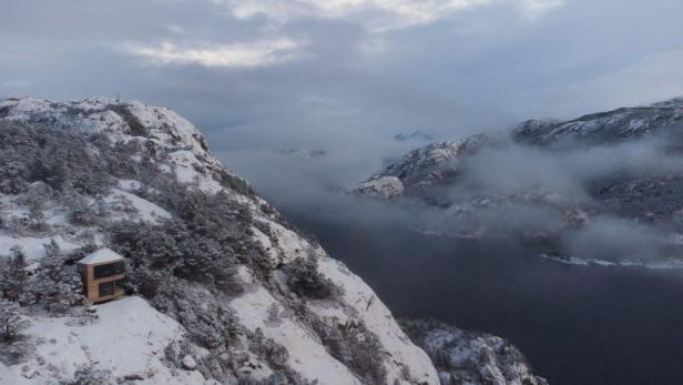 Eine kleine Holzhütte steht auf einem schneebedeckten Berghang mit Blick auf einen Fjord.