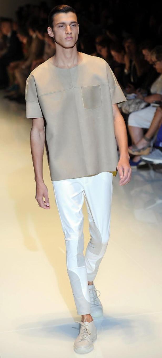 Ein Mann in beiger Kleidung und weißen Hosen auf einem Laufsteg.