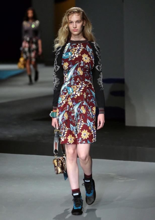 Eine Frau läuft auf einem Laufsteg und trägt ein geblümtes Kleid und Turnschuhe.