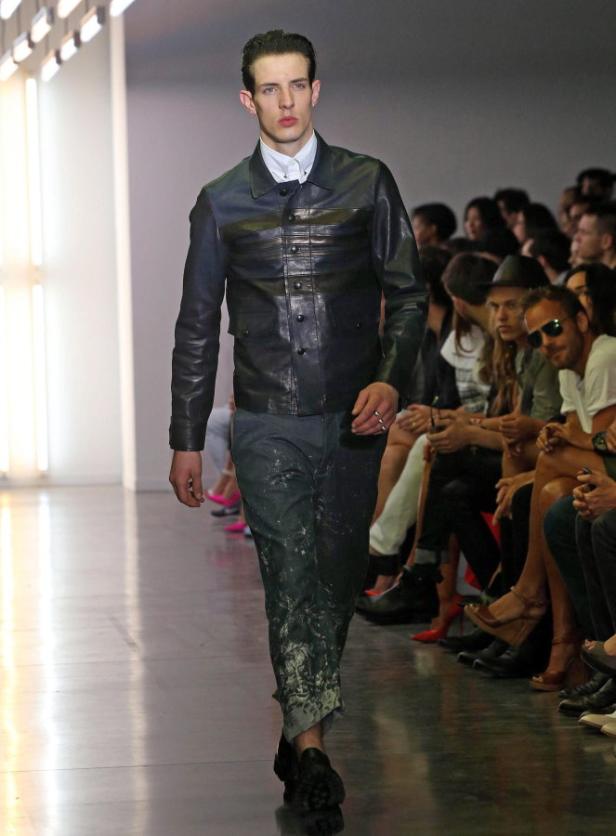 Ein Mann in Jacke und Hose läuft auf einem Laufsteg an Zuschauern vorbei.