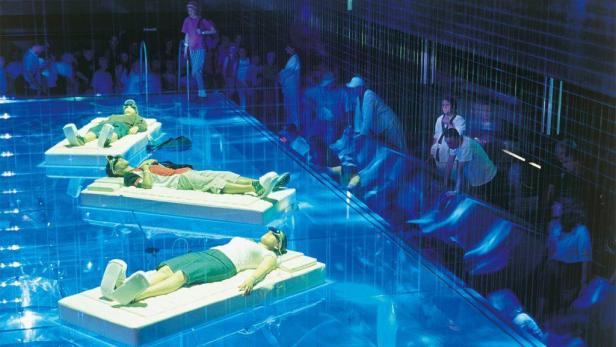 Menschen liegen auf Matratzen in einem blau beleuchteten Schwimmbad, während andere zusehen.