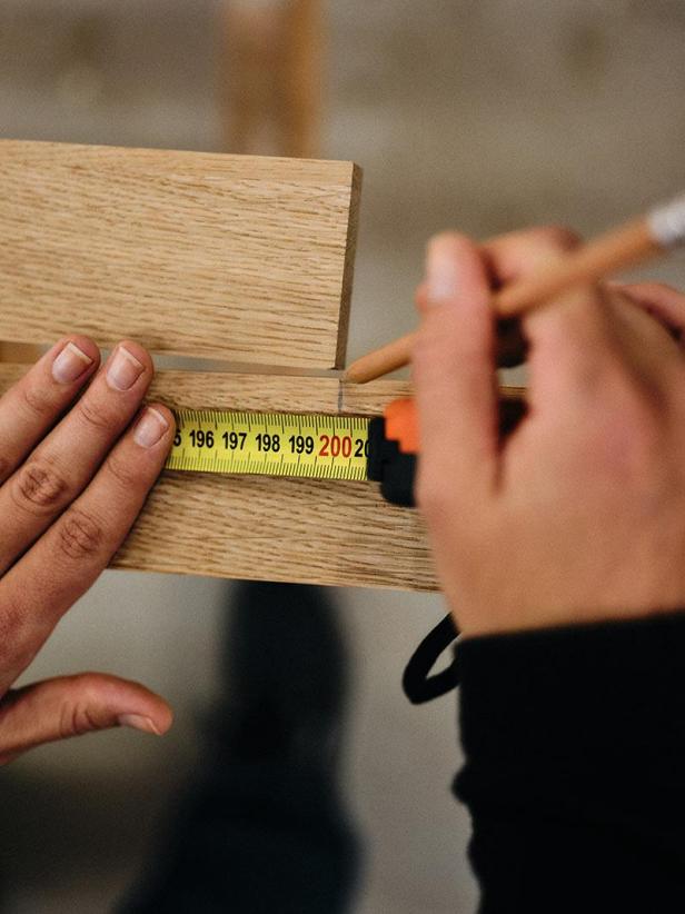 Eine Person misst ein Holzbrett mit einem gelben Maßband und markiert es mit einem Bleistift.