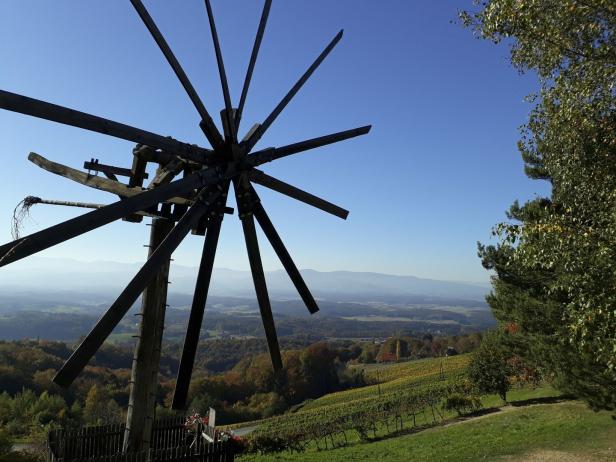 Eine hölzerne Windmühle überblickt eine Landschaft mit Weinbergen und bewaldeten Hügeln.