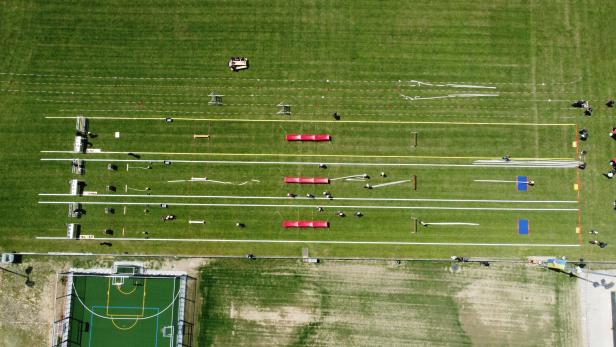 Eine Vogelperspektive zeigt ein grünes Feld mit mehreren Bahnen und Hindernissen, auf dem sich Personen bewegen.