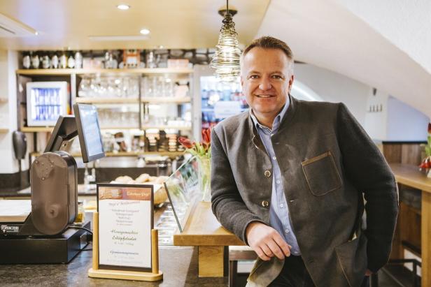 Ein lächelnder Mann in Trachtenjacke steht an der Theke eines Feinkostladens.